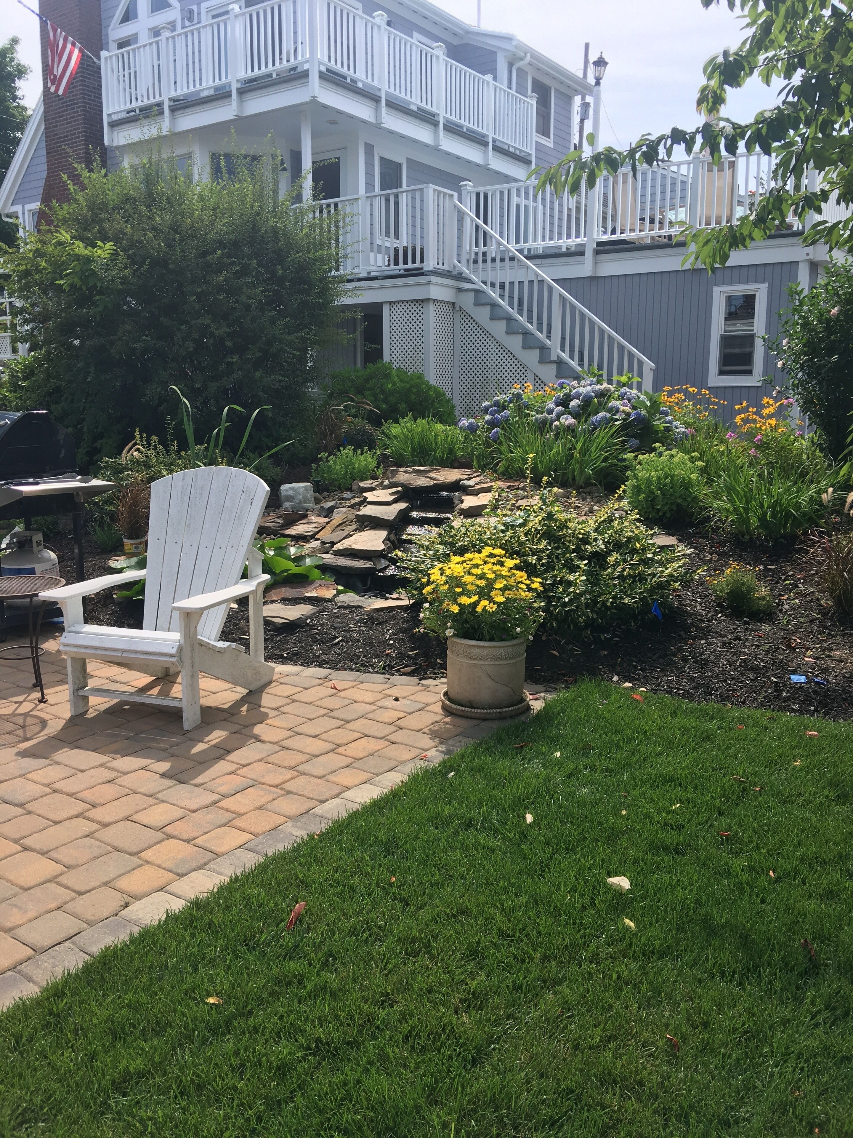 Une terrasse avec un fauteuil Adirondack blanc, un chemin de pierres et un jardin où poussent différentes plantes et fleurs.