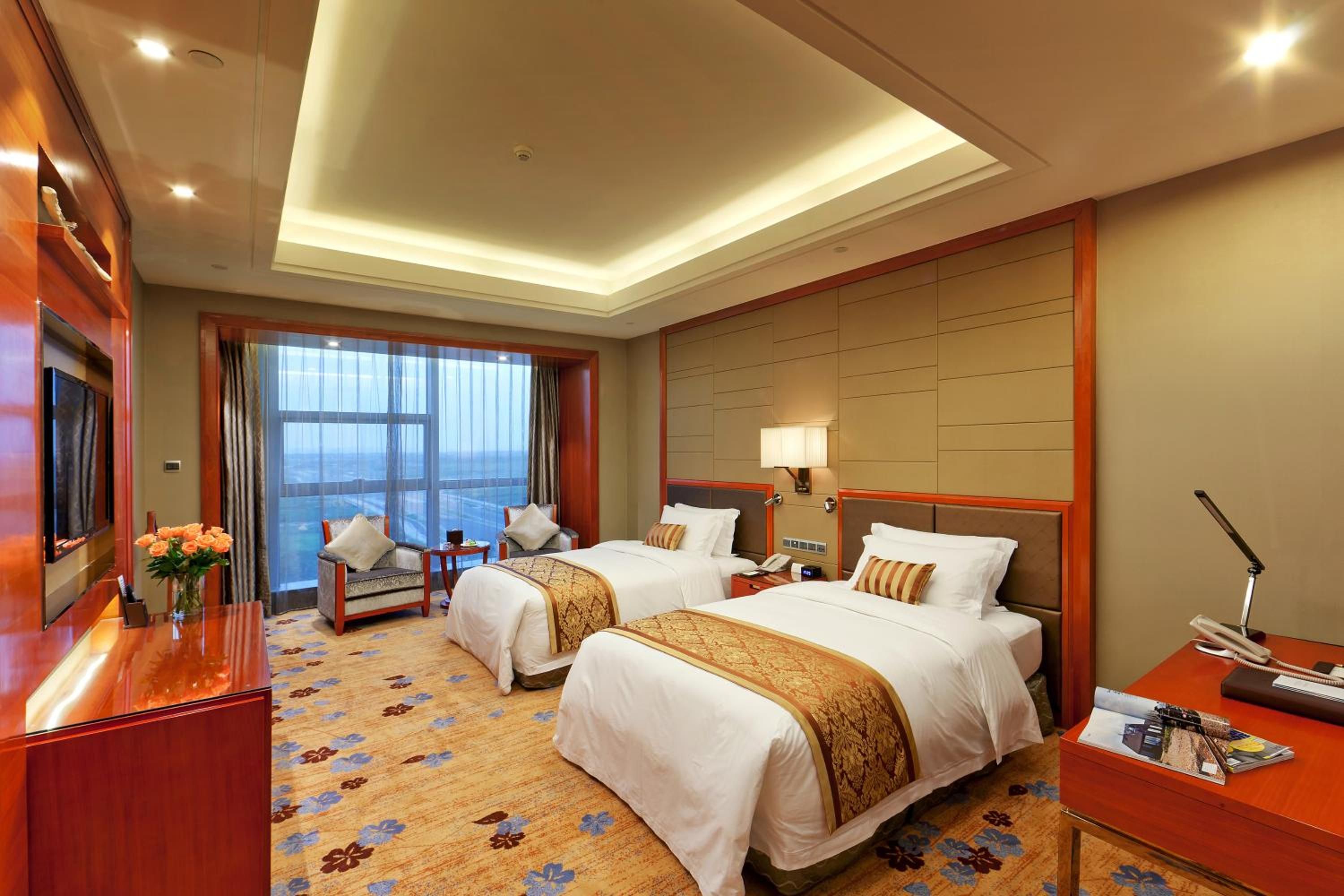 A hotel room with two beds, a desk, a chair, a TV, and a large window with curtains.