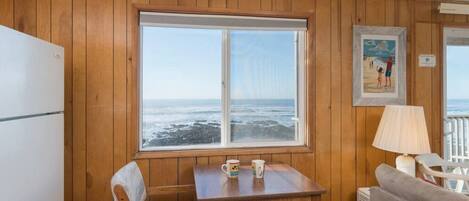 A room with wooden paneling, a large window overlooking the ocean, a table with chairs, a refrigerator, and a lamp.