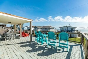 A wooden deck with turquoise chairs and a table, overlooking a beach with houses in the background.