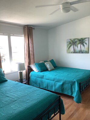 A room with two beds, a ceiling fan, a painting of palm trees, and a window with curtains.