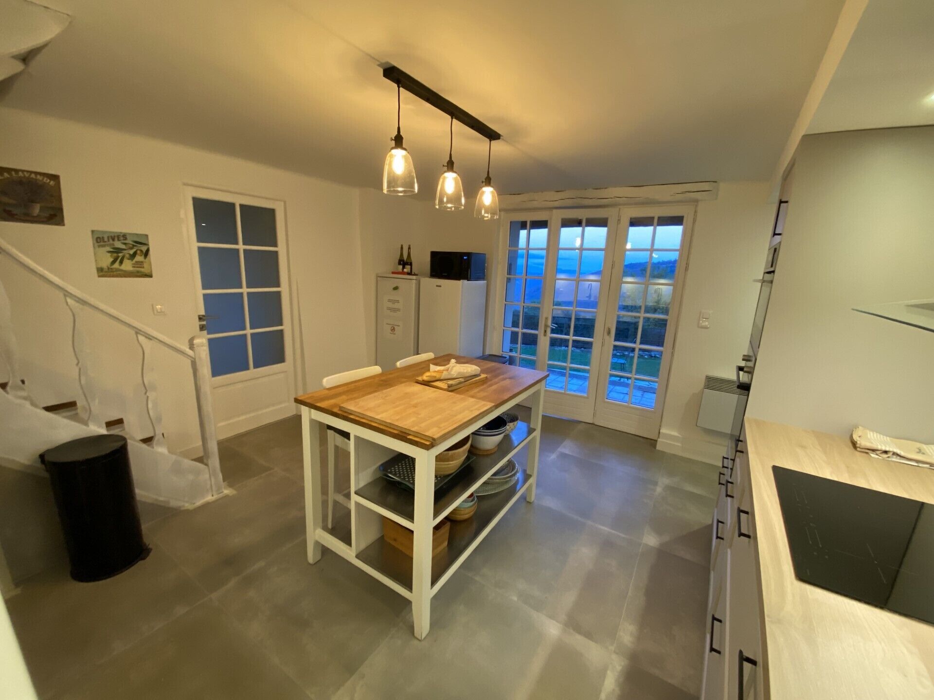 A modern kitchen with a wooden table, hanging pendant lights, and a view through glass doors to the outdoors.
