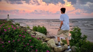 A man standing on rocks overlooking a body of water with a lighthouse in the distance.