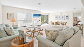 A spacious living area with a sofa, coffee table, and dining table. There is a TV, a lamp, and a window with blinds.
