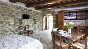 A rustic room with a stone wall, wooden ceiling beams, a dining table with chairs, a bed with a floral bedspread, and a kitchen area with a microwave and cabinets. - Agriturismo la Palazzetta di Assisi (Assisi)