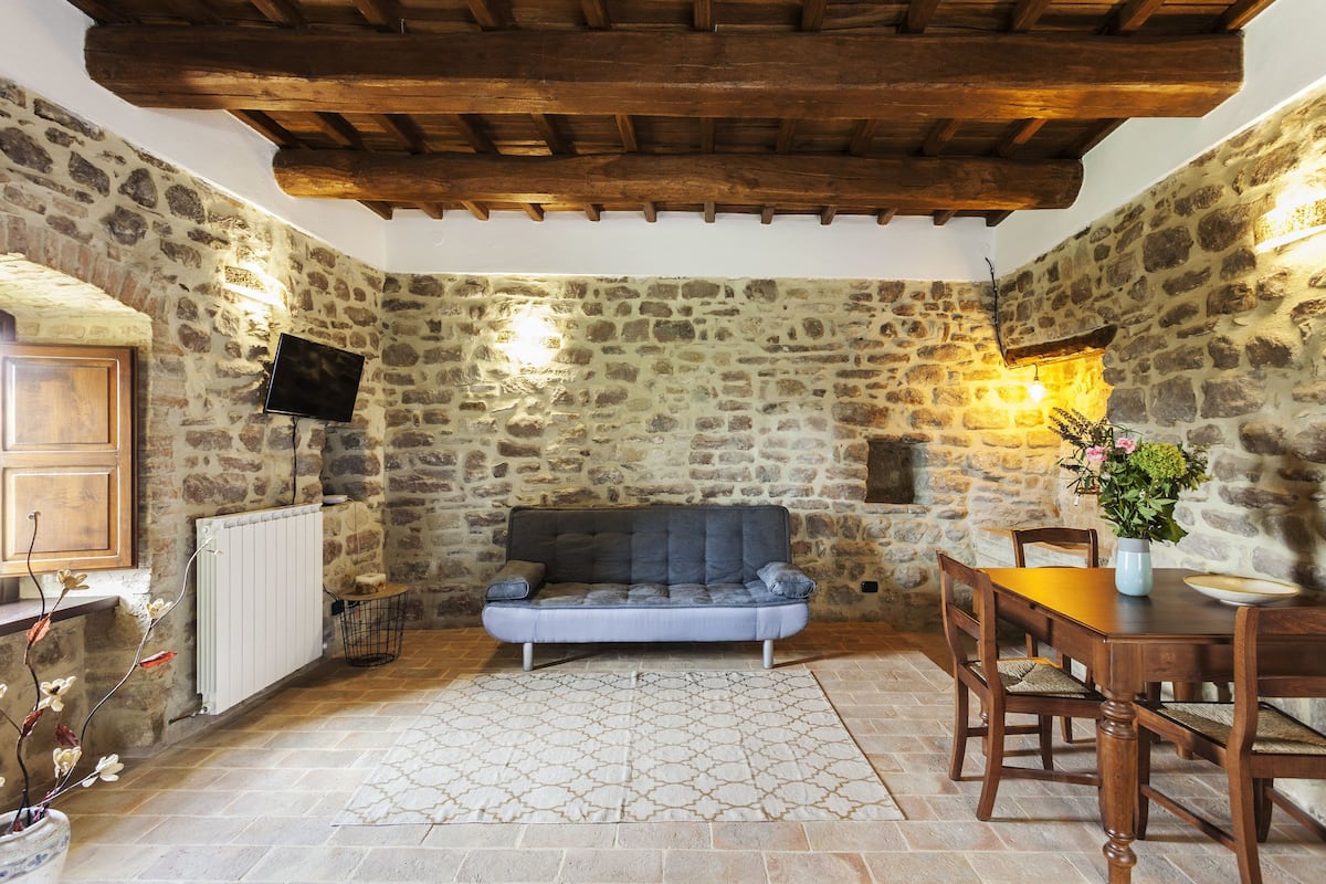 Une pièce rustique aux murs de pierre, avec un plafond en bois, un canapé gris, une table à manger avec des chaises et une plante en pot.