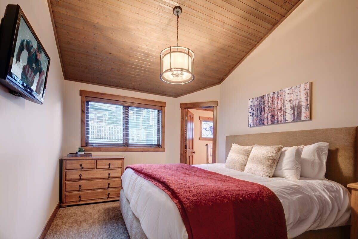Un dormitorio con techo de madera, una cama con ropa de cama blanca y roja, un aparador, un cuadro en la pared y una ventana con persianas.