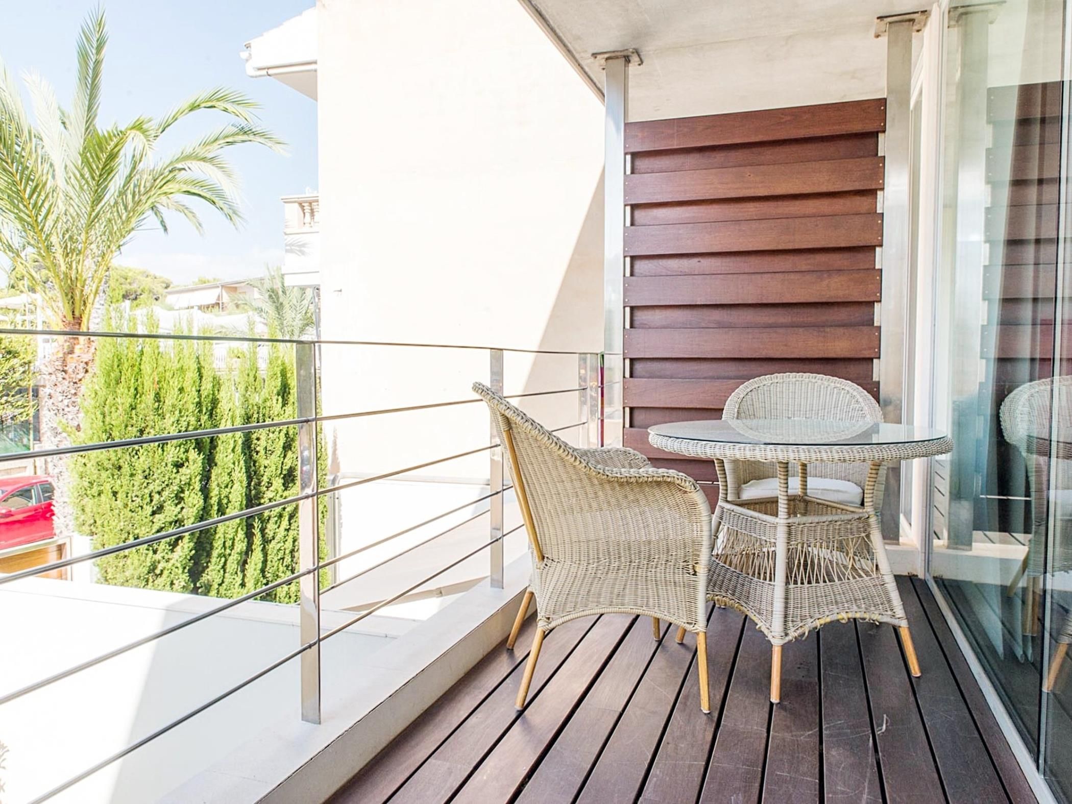 Een balkon met een tafel, stoelen, een glazen deur en uitzicht op groen en een gebouw.