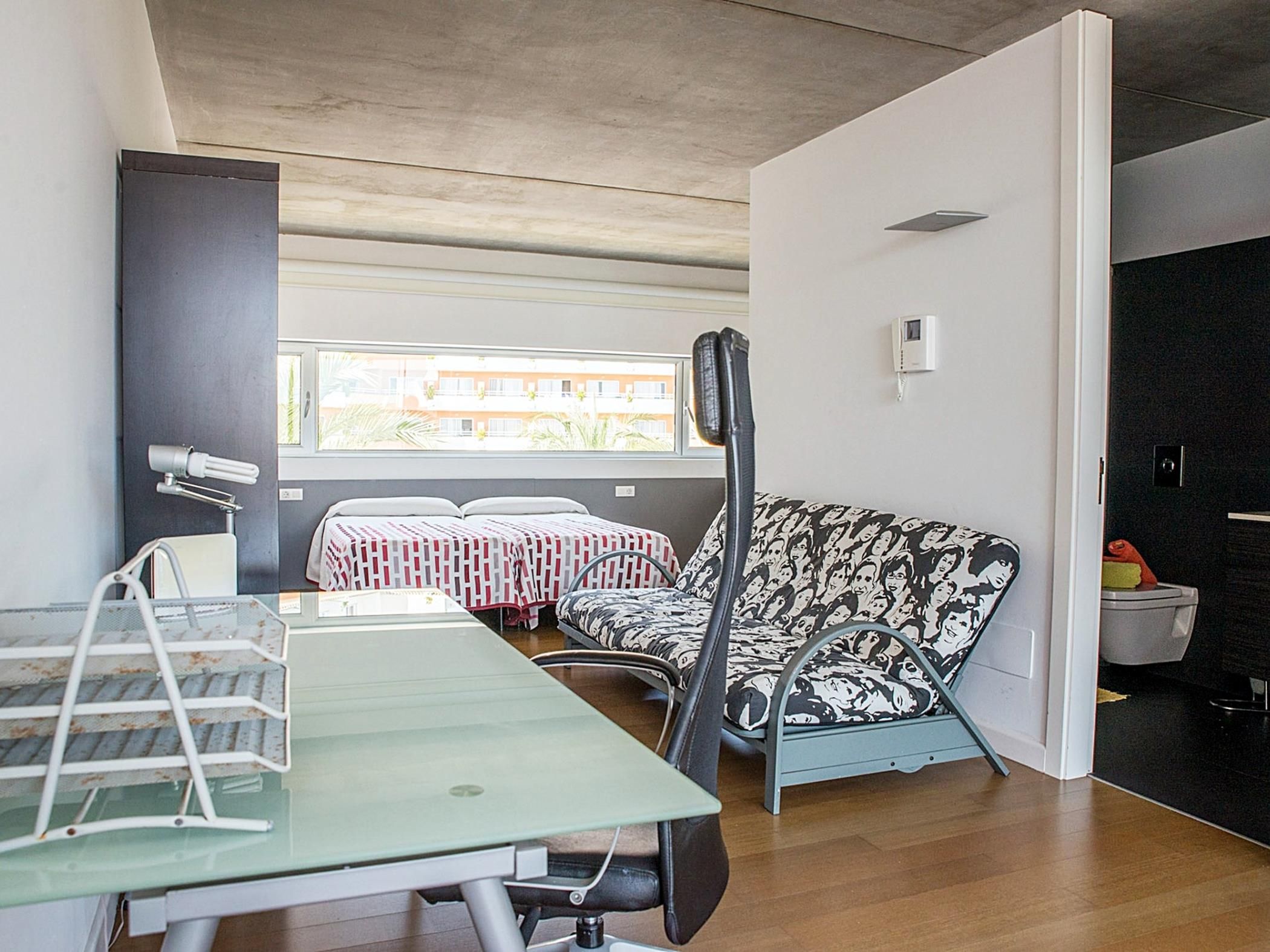 Un soggiorno moderno con un tavolo in vetro, un divano con rivestimento decorato, una finestra che si affaccia su edifici e una porta che dà su un angolo cottura.