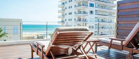Eine Holzterrasse mit zwei Liegestühlen, Blick auf das Meer und ein mehrgeschossiges Gebäude im Hintergrund.