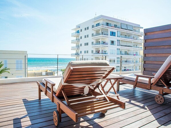 A wooden deck with two lounge chairs, a view of the ocean, and a multi-story building in the background.