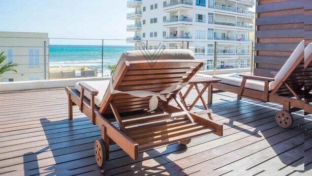 A wooden deck with two lounge chairs and a small table, overlooking a beach and a multi-story building with balconies.