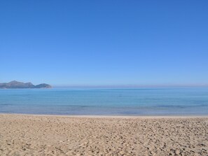 A sandy beach with clear blue water and distant mountains.