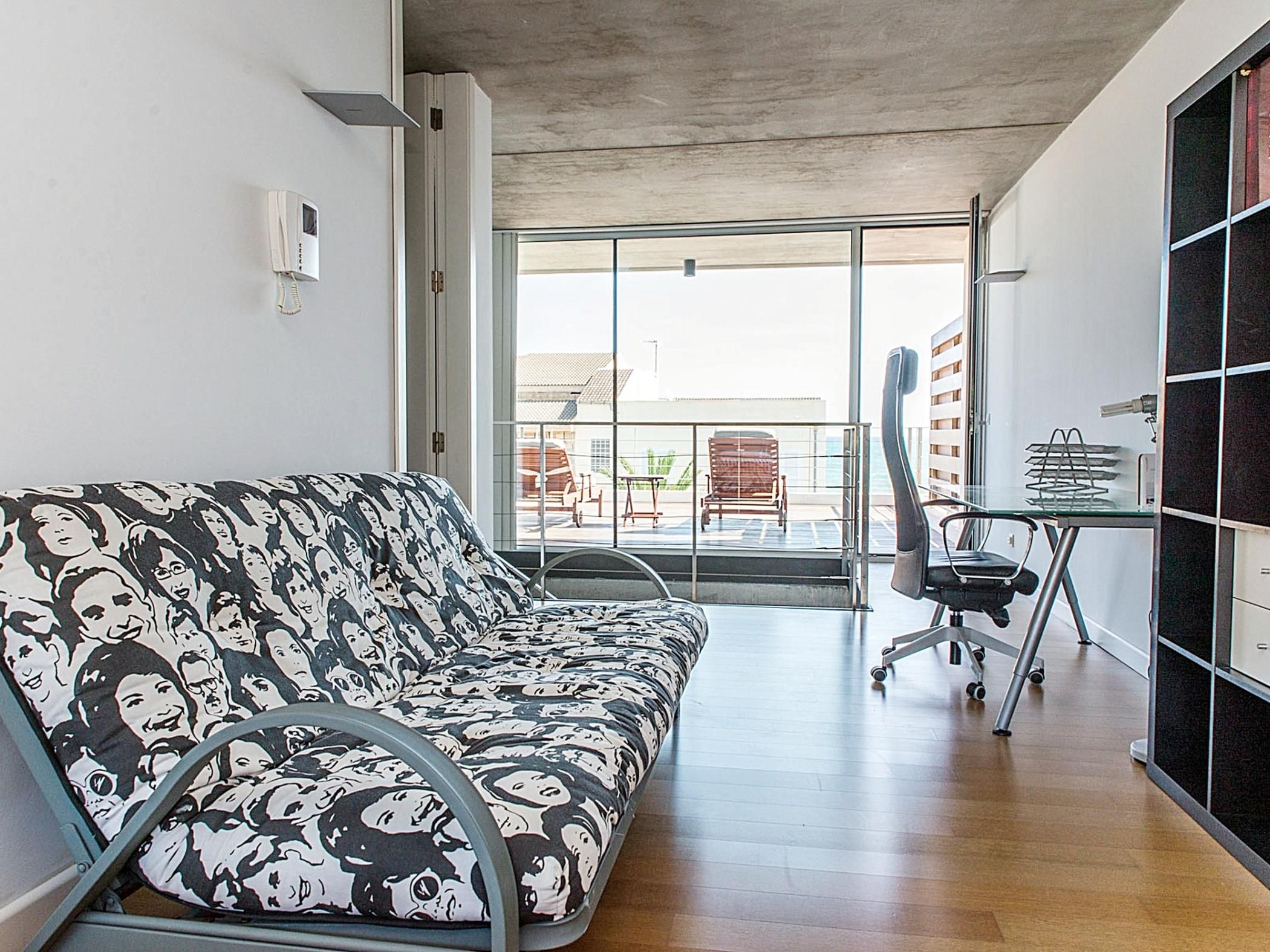 A modern living room with a patterned sofa, a dining area with a chair and table, and a large glass door leading to an outdoor balcony.