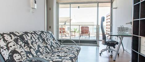 A modern living room with a patterned sofa, a dining area with a chair and table, and a large glass door leading to an outdoor balcony.