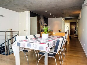 En spiseplads med et bord dækket til seks personer, omgivet af stole, en potteplante og et køkken i baggrunden.