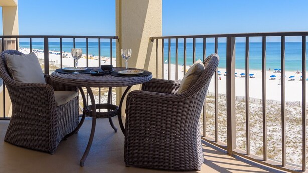 A balcony with wicker furniture, a table, and a glass of wine. The view includes a beach with umbrellas and people. -  Stunning Beachfront View Condo (Orange Beach)