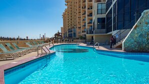 A swimming pool with lounge chairs and a stone wall, surrounded by a high-rise building. -  Stunning Beachfront View Condo (Orange Beach)