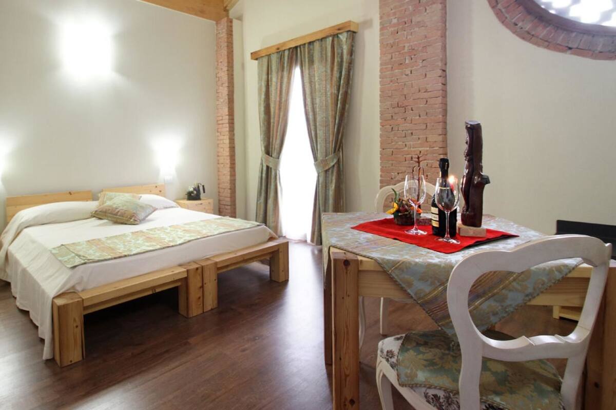 A bedroom with a wooden bed, a small bedside table, a brick wall, and a dining area with a table and chairs.