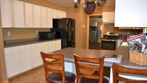 A kitchen with white cabinets, a black refrigerator, a microwave, and a stove. There are three wooden chairs with black cushions. A magazine rack with a 'Rapid City Visitors Guide' is visible. - Keystone Crossing (Keystone)