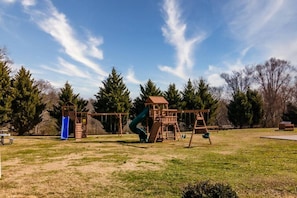 Kids can play on the playground's slide and swings in a lush green field surrounded by trees.