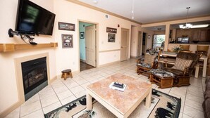 A living room with a fireplace, television, and a ceiling fan. There are framed pictures on the wall and a dining area with chairs. - The Cove - Unit 1302 - FREE Attraction Ticket with EACH night Stay Including Dollywood (Pigeon Forge)