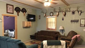 A living room with a brown couch, blue armchair, and a wooden chair with a blanket. There are mounted animal skulls on the wall, a television, and a ceiling fan. - Sportsman's Paradise (Stockton)