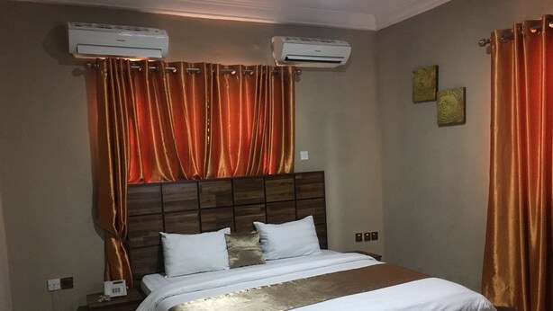 A hotel room with a large bed, two air conditioning units, a wooden headboard, and a brown accent on the bedspread. - Momak 4 Hotel & Suites (Ibadan)