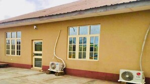A building with a beige exterior, a tiled roof, and two windows. There are two air conditioning units on the ground. - Momak 4 Hotel & Suites (Ibadan)