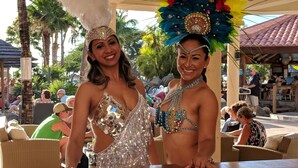 Deux personnes, vêtues de costumes élaborés, se trouvent dans un décor extérieur aux allures tropicales, avec des palmiers et des espaces aménagés pour s’asseoir.
