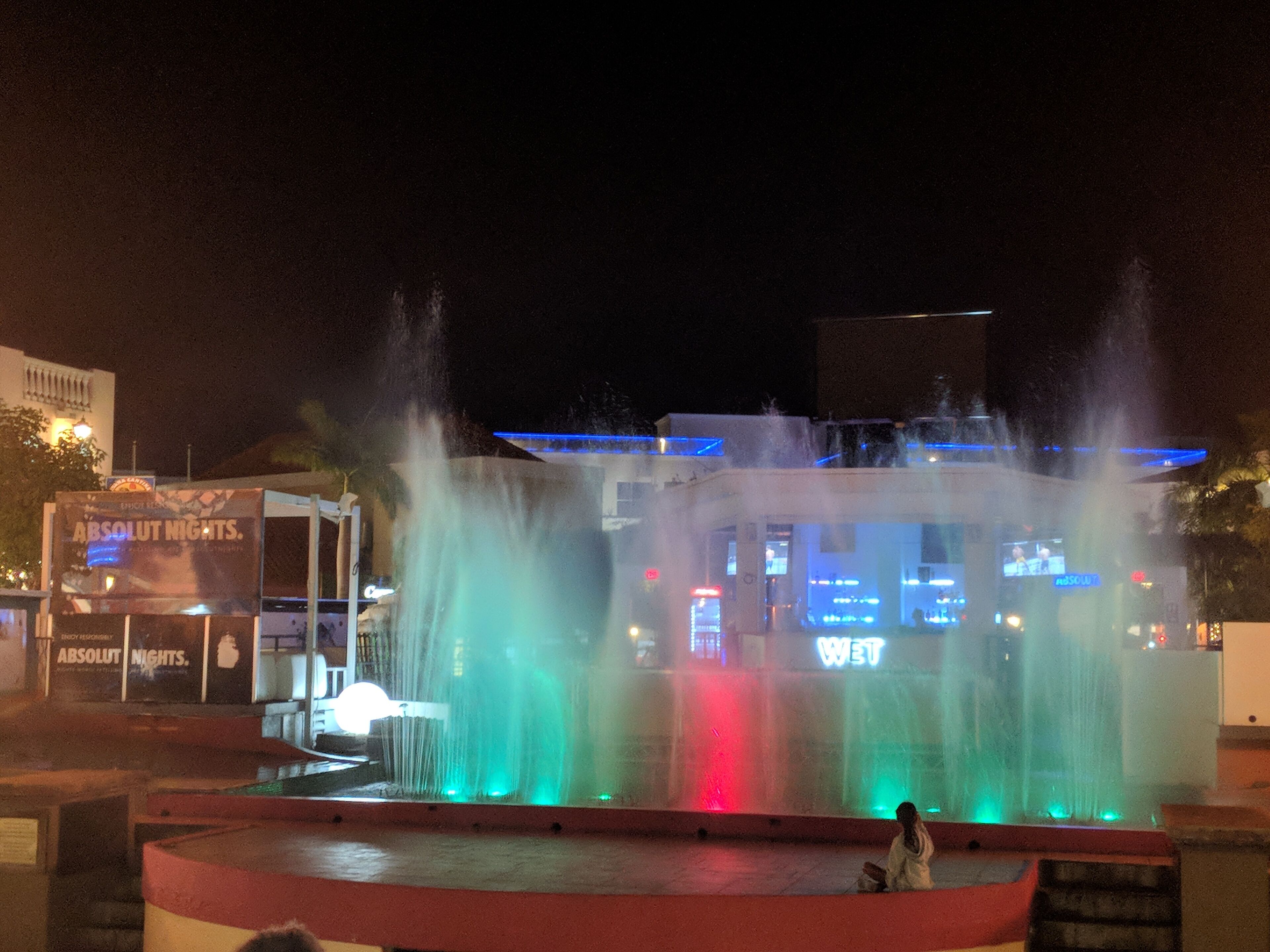 Eine Nachttasse mit einem Springbrunnen auf einem öffentlichen Platz, beleuchtet von bunten Lichtern.