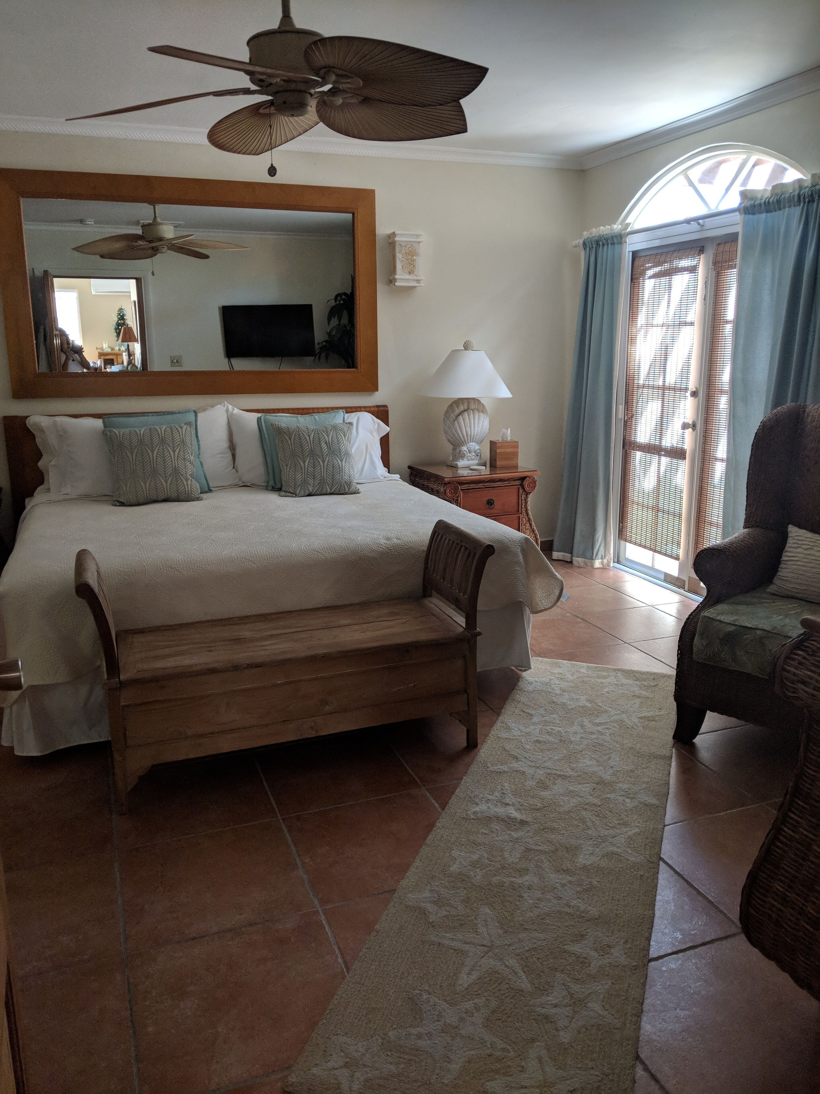 A bedroom with a large bed, a wooden bench, a ceiling fan, a nightstand with a lamp, and a chair.