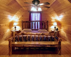 Une chambre dans un chalet en rondins, avec un lit en bois, deux lampes de chevet, un ventilateur de plafond et une fenêtre ornée de rideaux violets.