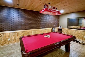 Une table de billard recouverte d’un feutre rouge, avec un cadre en bois, et un parasol rouge et blanc à l’effigie de la marque Budweiser placé au-dessus.