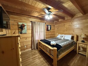 A wooden cabin bedroom with a bed, a nightstand, a ceiling fan, and a window.