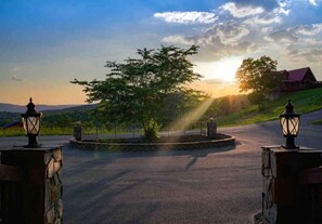 A paved driveway leading to a gated area with a tree in the center, surrounded by stone walls and two lantern-style lights.