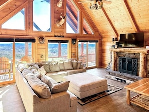 A log cabin interior with a large sectional sofa, a fireplace, and a view through the large windows. - 8 BR Mountaintop Lodge with views - Sleeps 22 (Dobson)