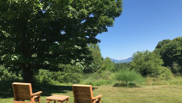 Zwei Holzstühle und ein kleiner Tisch stehen auf einer Wiese mit einem großen Baum und einem Berg im Hintergrund.