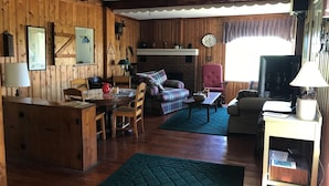 Ein gemütliches Blockhaushaus-Interieur mit Holzwänden, einem Essbereich, einem Wohnzimmer mit Sofa und Sesseln sowie einem Fernseher.