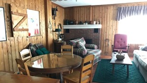 Ein gemütliches Wohnzimmer mit einem runden Holztisch, einer Couch und einem Sessel. Ein Kamin mit Sims und verschiedenen Dekorationselementen ist vorhanden.
