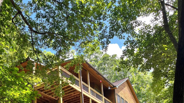 A two-story wooden cabin with a balcony on each floor, surrounded by trees and a grassy area.