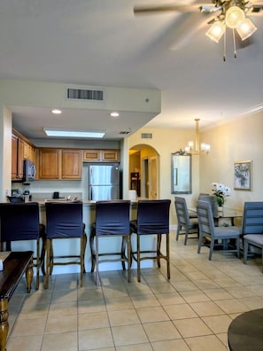 A modern kitchen with a dining area, including a bar with chairs, a dining table with chairs, and a ceiling fan.