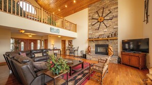 A spacious living room with a large sectional sofa, a fireplace, a TV, and a balcony with wooden railings. - Scenic Mountaintop Chalet w/ Hot Tub & Fire Pit (McHenry)