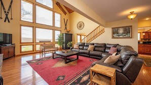 A spacious living room with a leather sofa, a coffee table, and a red rug. There are large windows, a staircase, and a painting on the wall. - Scenic Mountaintop Chalet w/ Hot Tub & Fire Pit (McHenry)