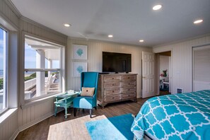 Un dormitorio con un ventanal grande, una cama con colcha azul estampada, un aparador de madera, un sillón azul turquesa y una mesita con libros.