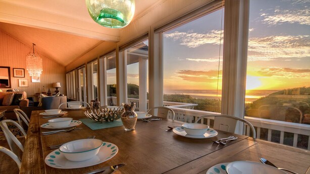 A dining area with a long table set for a meal, large windows offering a view of the sunset, and a chandelier hanging from the ceiling. - Beach Front Panoramic Vistas! Miles of Sandy Beach! Hot Tub! Dog Friendly! (Waldport)