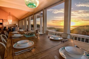 A dining area with a long table set for a meal, large windows offering a view of the sunset, and a chandelier hanging from the ceiling.