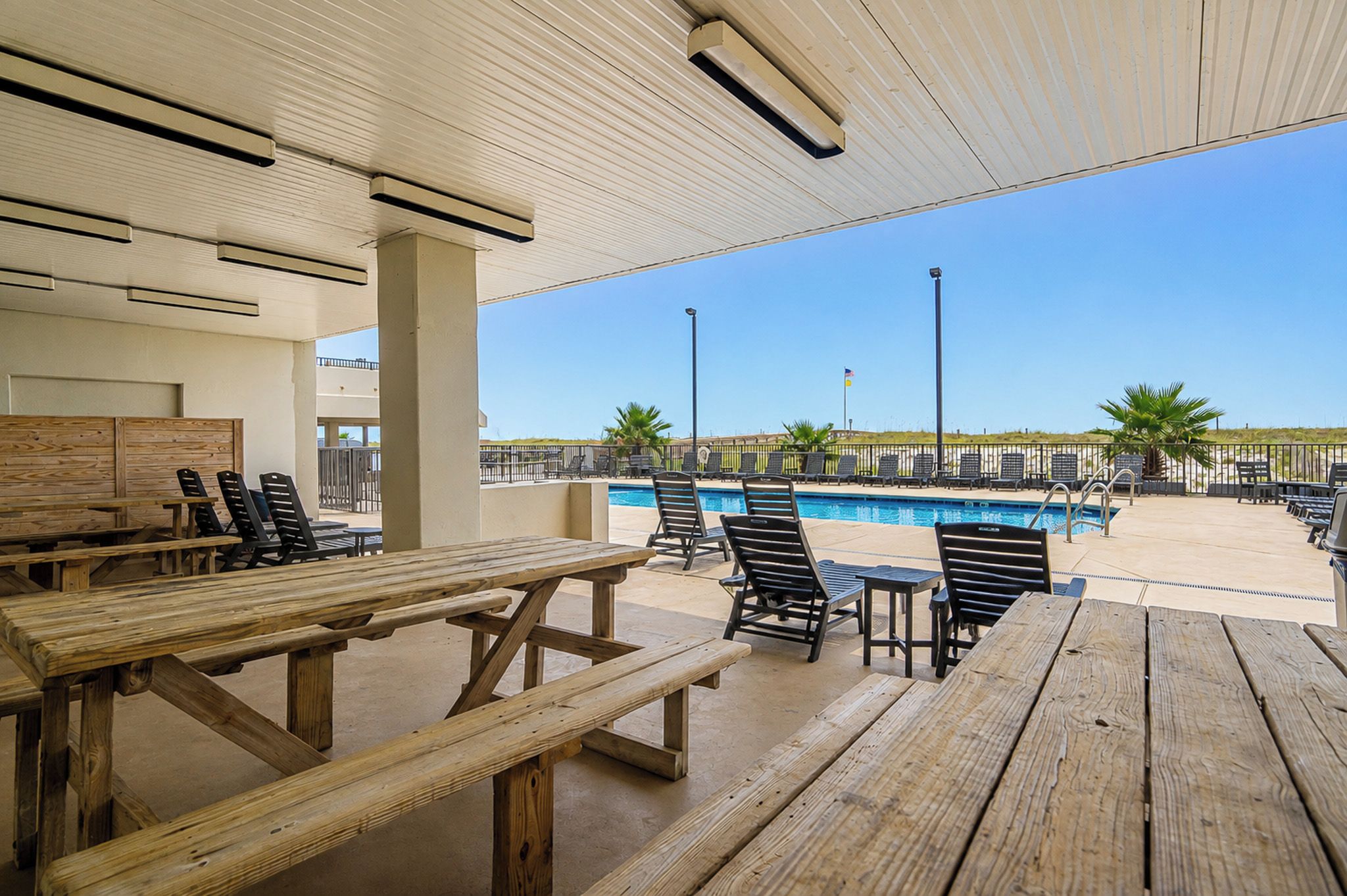 An outdoor pool area with wooden picnic tables and lounge chairs, a clear sky, and a view of the beach.