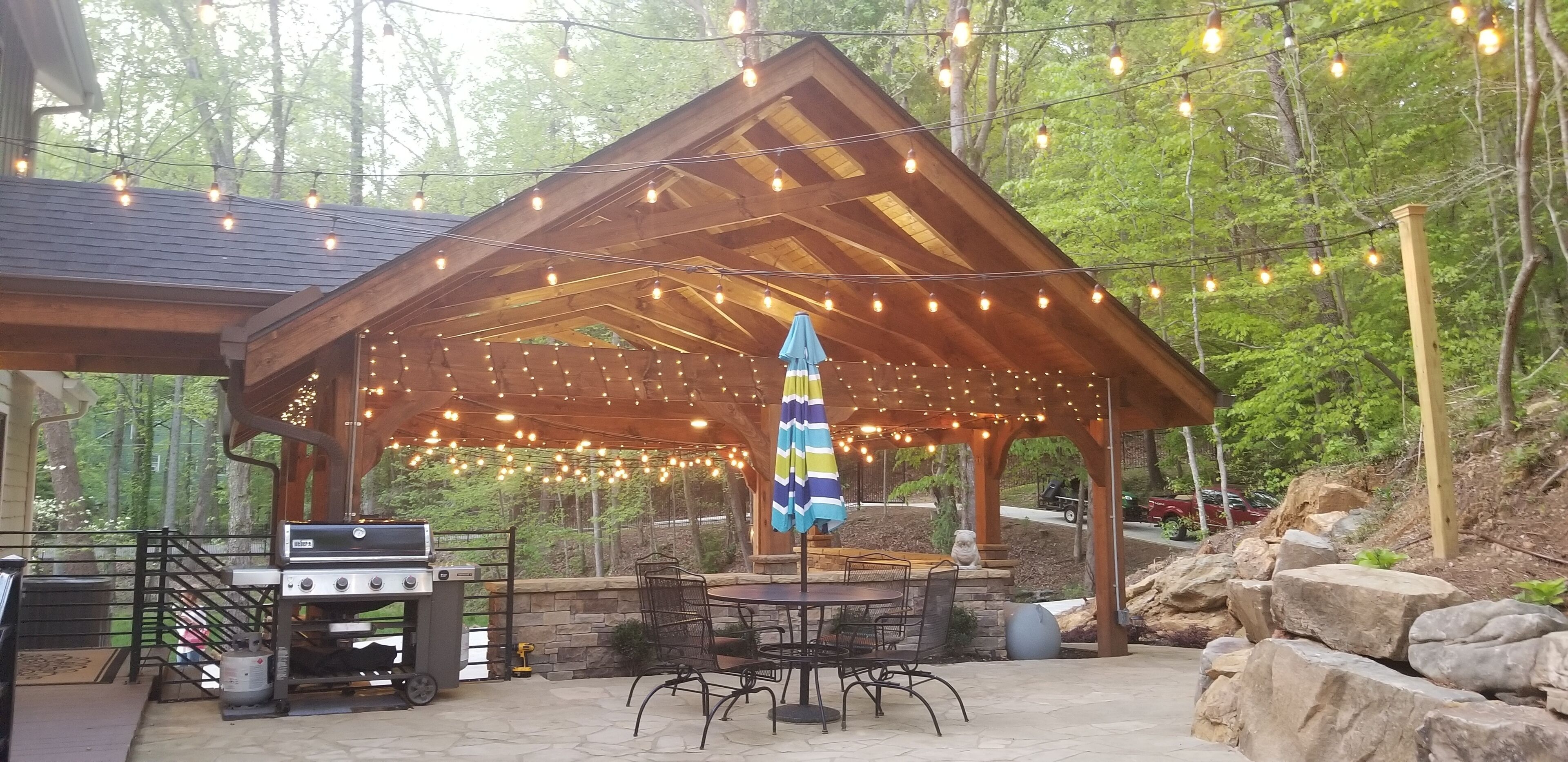 Un patio exterior con una pérgola de madera, luces colgantes, una parrilla y asientos de piedra.