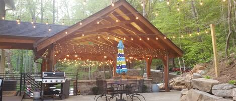 Un patio exterior con una pérgola de madera, luces colgantes, una parrilla y asientos de piedra.